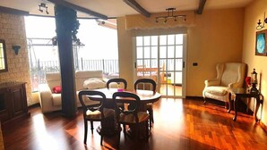 A spacious living room with a wooden floor, a dining area with a table and chairs, a sofa, and a chair. There is a balcony with a railing and a view outside. - Adeje Tijoco Alto Overseas Real Estate Villa (Palm-Mar)