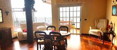 A spacious living room with a wooden floor, a dining area with a table and chairs, a sofa, and a chair. There is a balcony with a railing and a view outside.