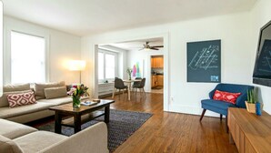 Een moderne woonkamer met een bank, salontafel en een blauwe fauteuil. Er is een eethoek met een tafel en stoelen, en een keuken op de achtergrond.