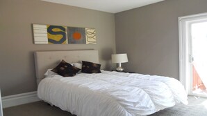Un dormitorio con cama, almohadas, mesita de noche, lámpara y una obra de arte en la pared.