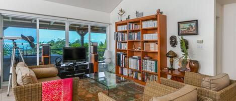 Un salon moderne avec une table basse en verre, des meubles en osier, une bibliothèque et une vue sur l’extérieur.