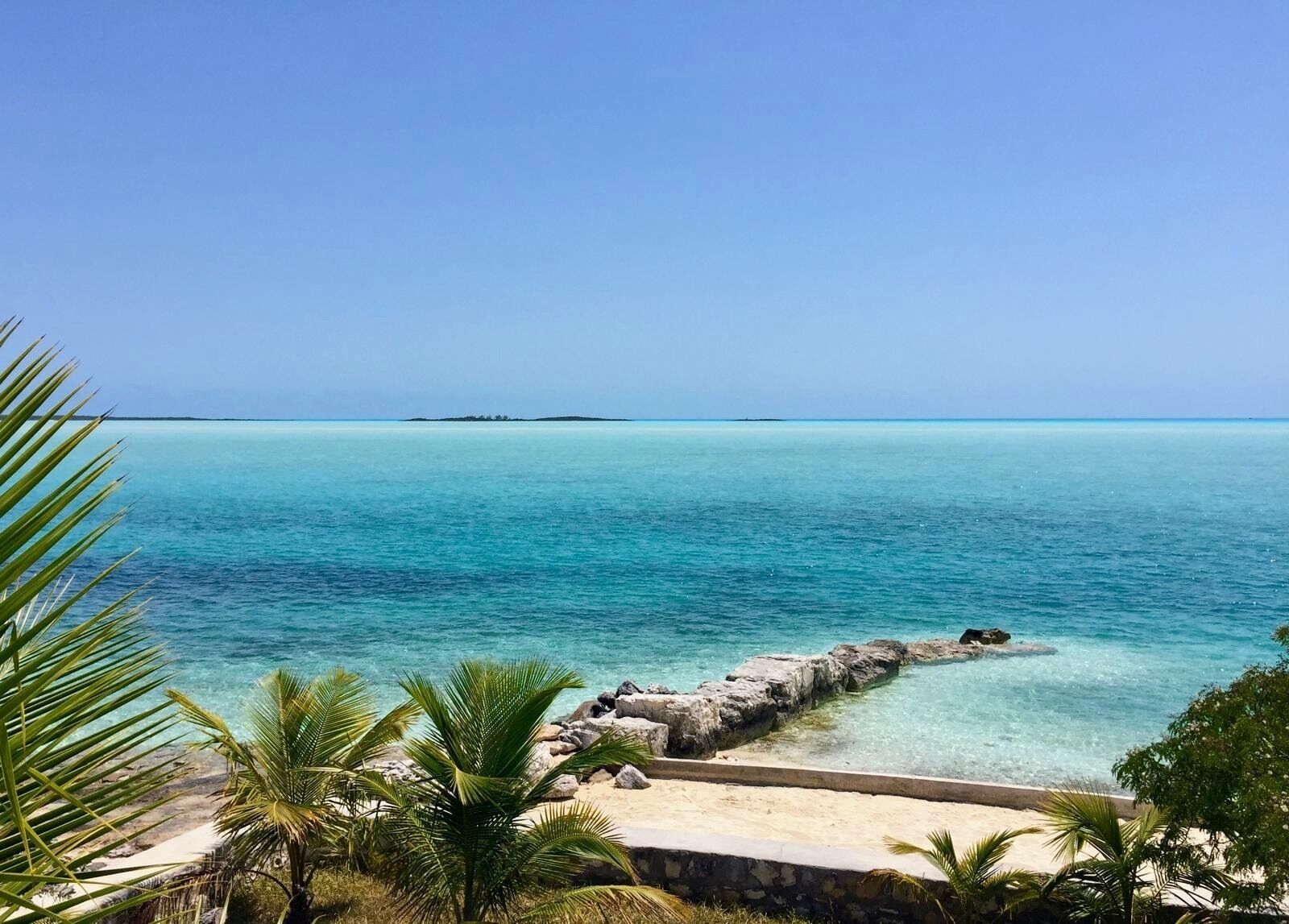 A clear turquoise sea with a rocky breakwater, palm trees, and a distant island.
