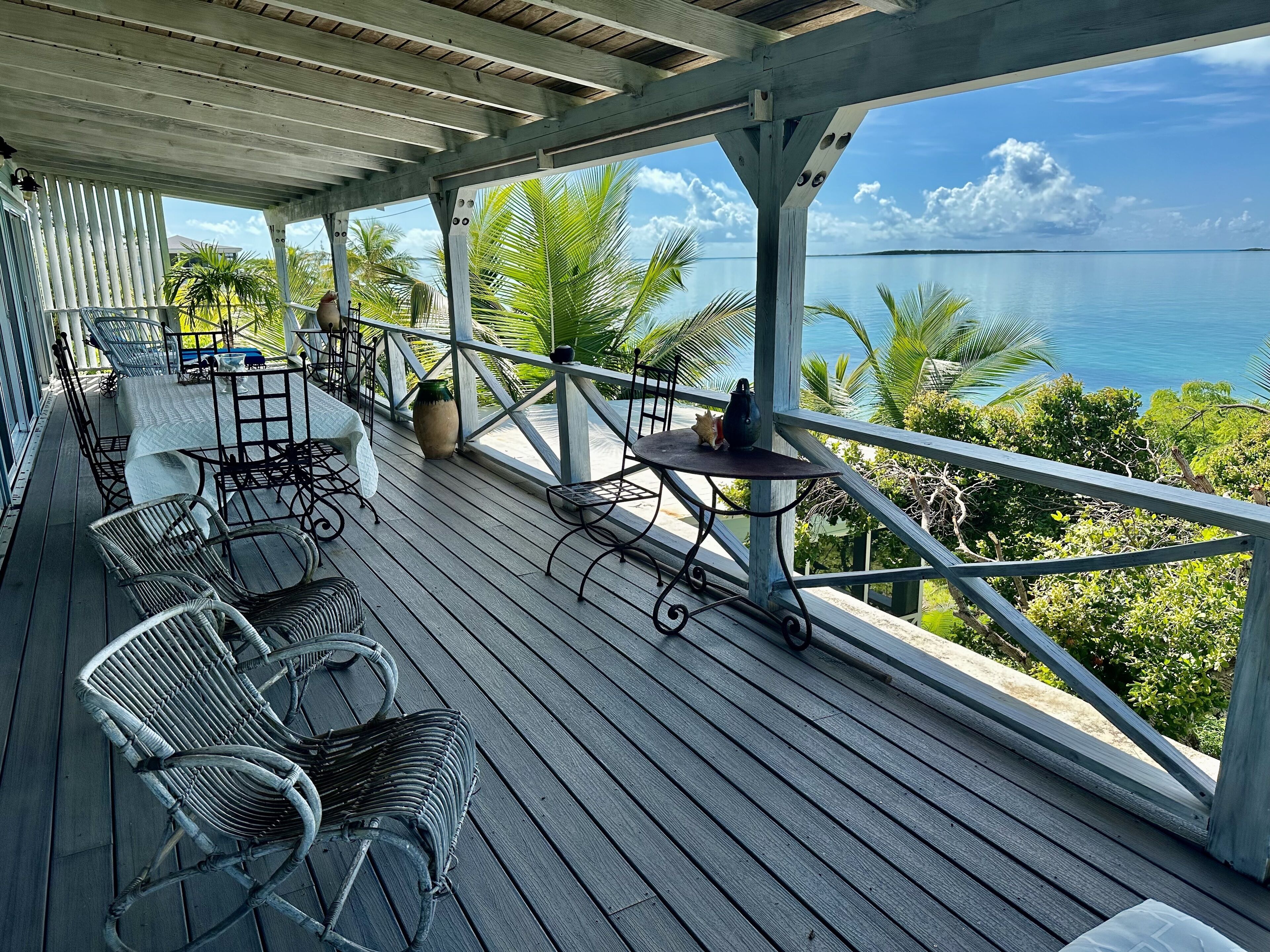 A wooden deck with outdoor furniture, a table, and chairs, overlooking a body of water and palm trees.