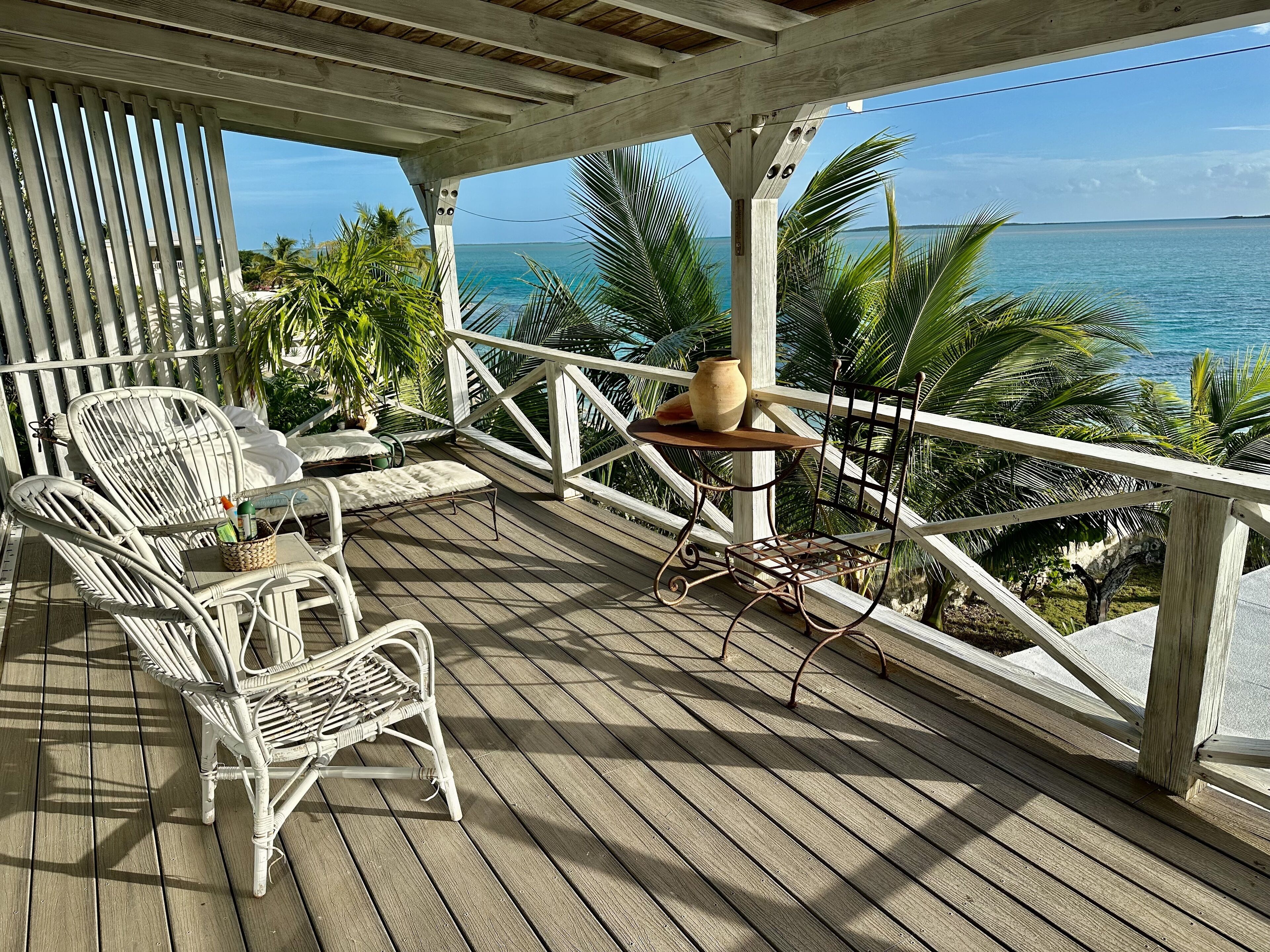 A wooden deck with white wicker chairs and a metal table, overlooking a clear blue ocean.