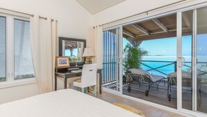 A bedroom with a bed, a desk with a chair, a window with curtains, and a balcony with a view of the ocean. - Bonefish Bella Vista - Beachfront Retreat, Tropical Garden & Natural Ocean Pool (Georgetown)