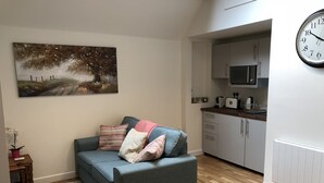 A living room with a grey sofa, a painting of a landscape, and a kitchen area with cabinets and appliances. - Perfect for walkers, Goodwood, National Trust, Short Stay (Bramshott Chase)