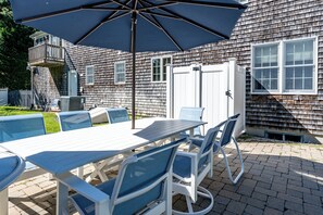 An outdoor patio with a table and chairs, a large umbrella, and a house with a shingled exterior.