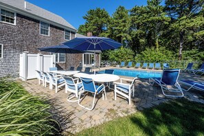 An outdoor pool area with lounge chairs and a table, surrounded by a brick patio and a wooden fence.