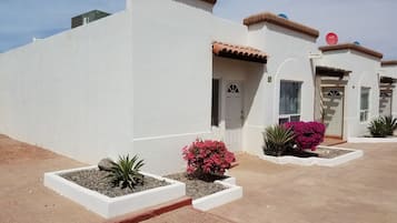 A row of white buildings with terracotta roofs, each with a door and windows. There are flower beds with pink flowers and a small plant in front of each building.