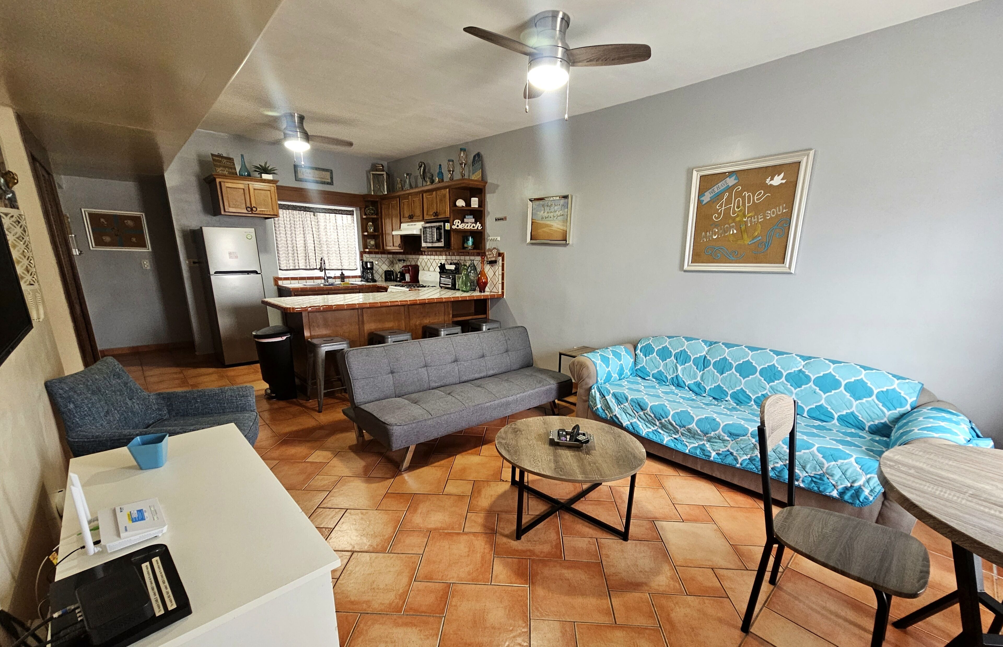 Un espacio habitable compacto con kitchenette, un sofá gris, un sofá azul con estampado, una mesa de centro redonda y una mesa de comedor con sillas.