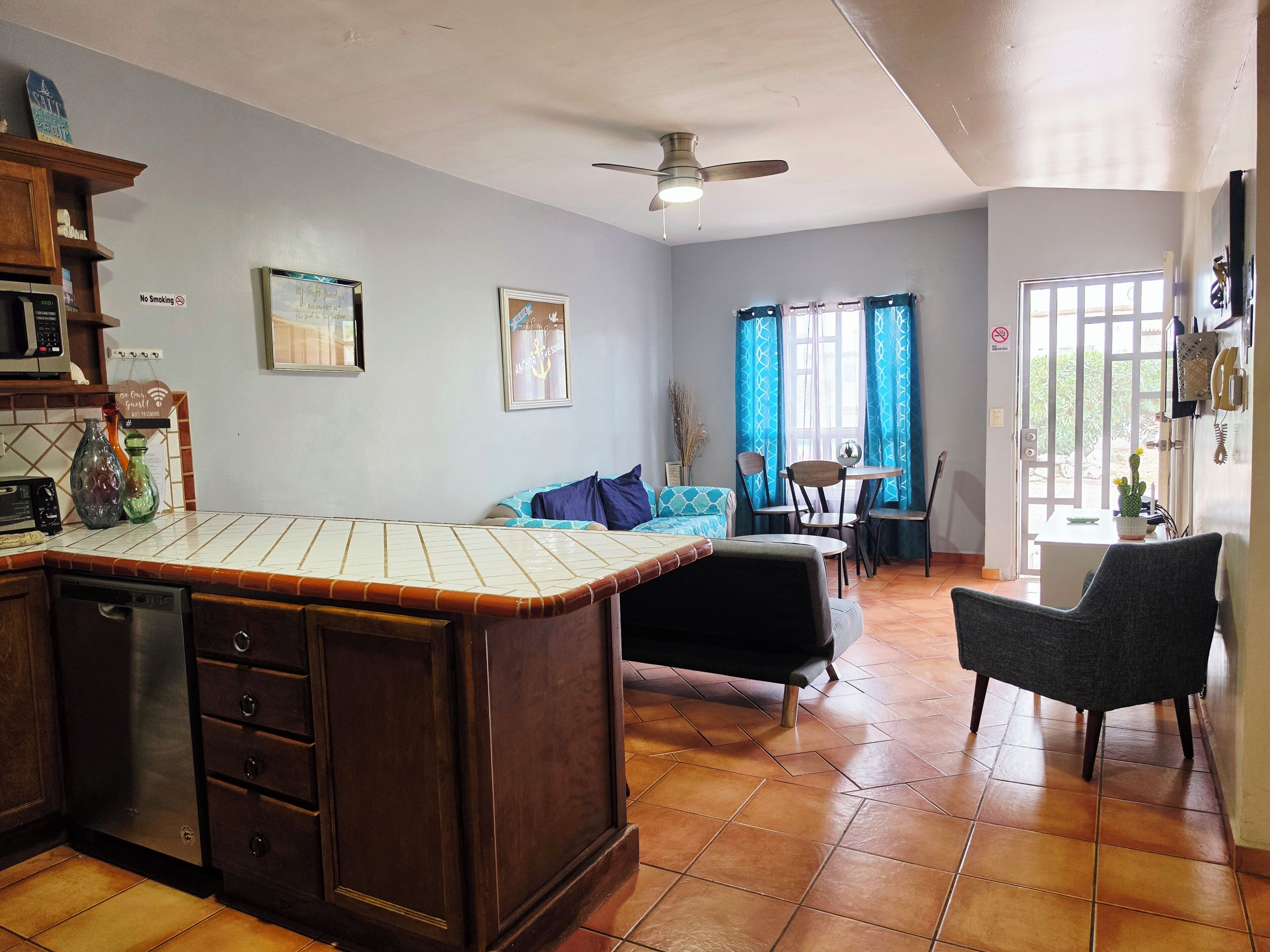 Uma cozinha com uma ilha central, área de jantar com cadeiras e mesa, ventilador de teto e vista pela janela com cortinas.