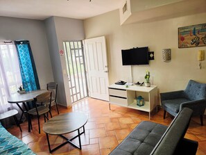 A living room with a sofa, a dining table with chairs, a TV mounted on the wall, and a door leading outside.