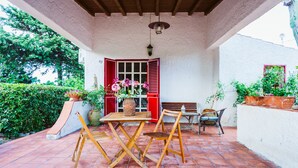 Een overdekt terras met een houten tafel en stoelen, een bankje en plantenpotten.