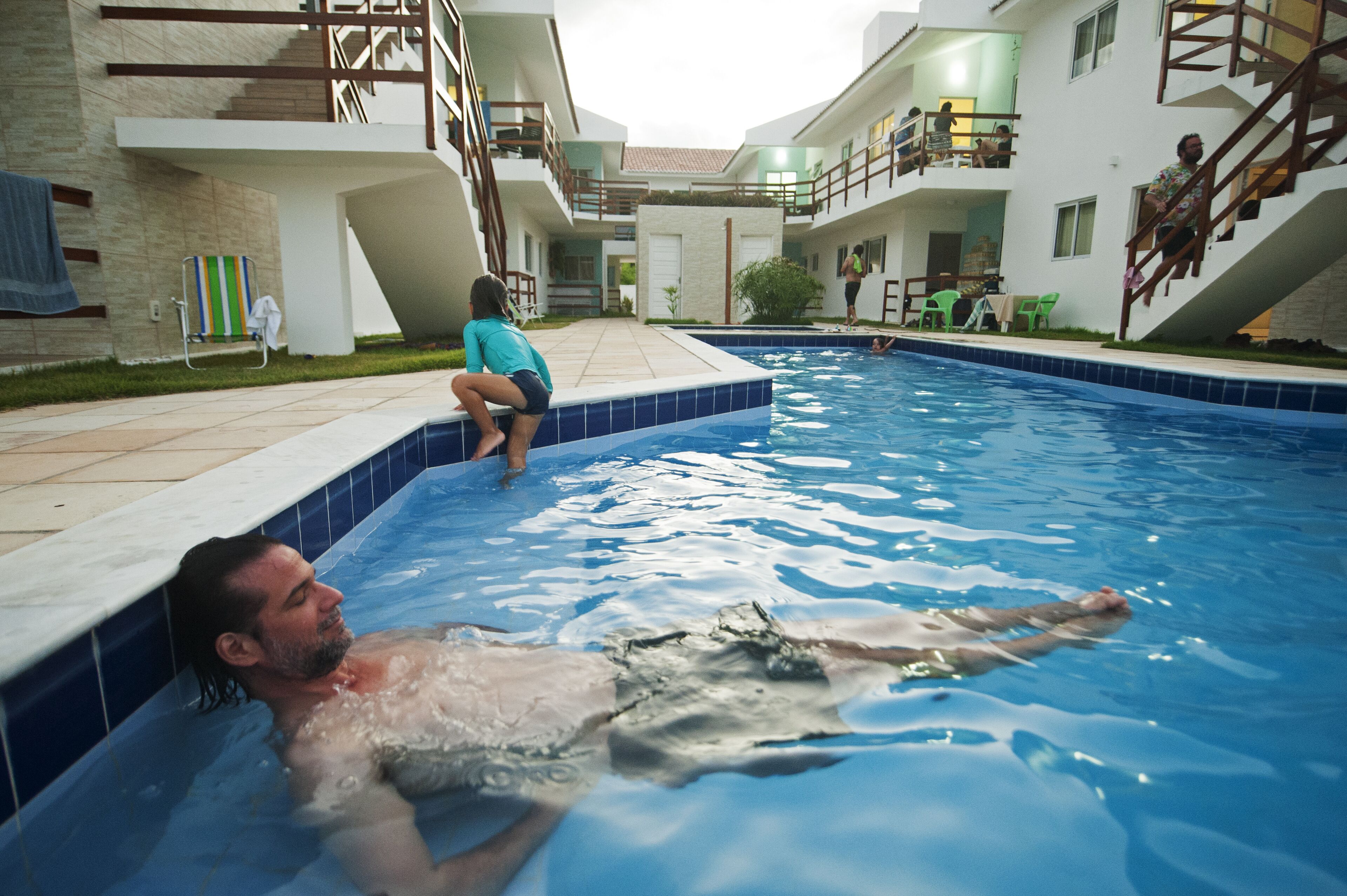 Uma área de piscina com uma pessoa deitada na água, outra sentada na borda e uma escadaria que leva a um nível superior.