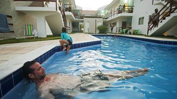 Uma área de piscina com uma pessoa deitada na água, outra sentada na borda e uma escadaria que leva a um nível superior.
