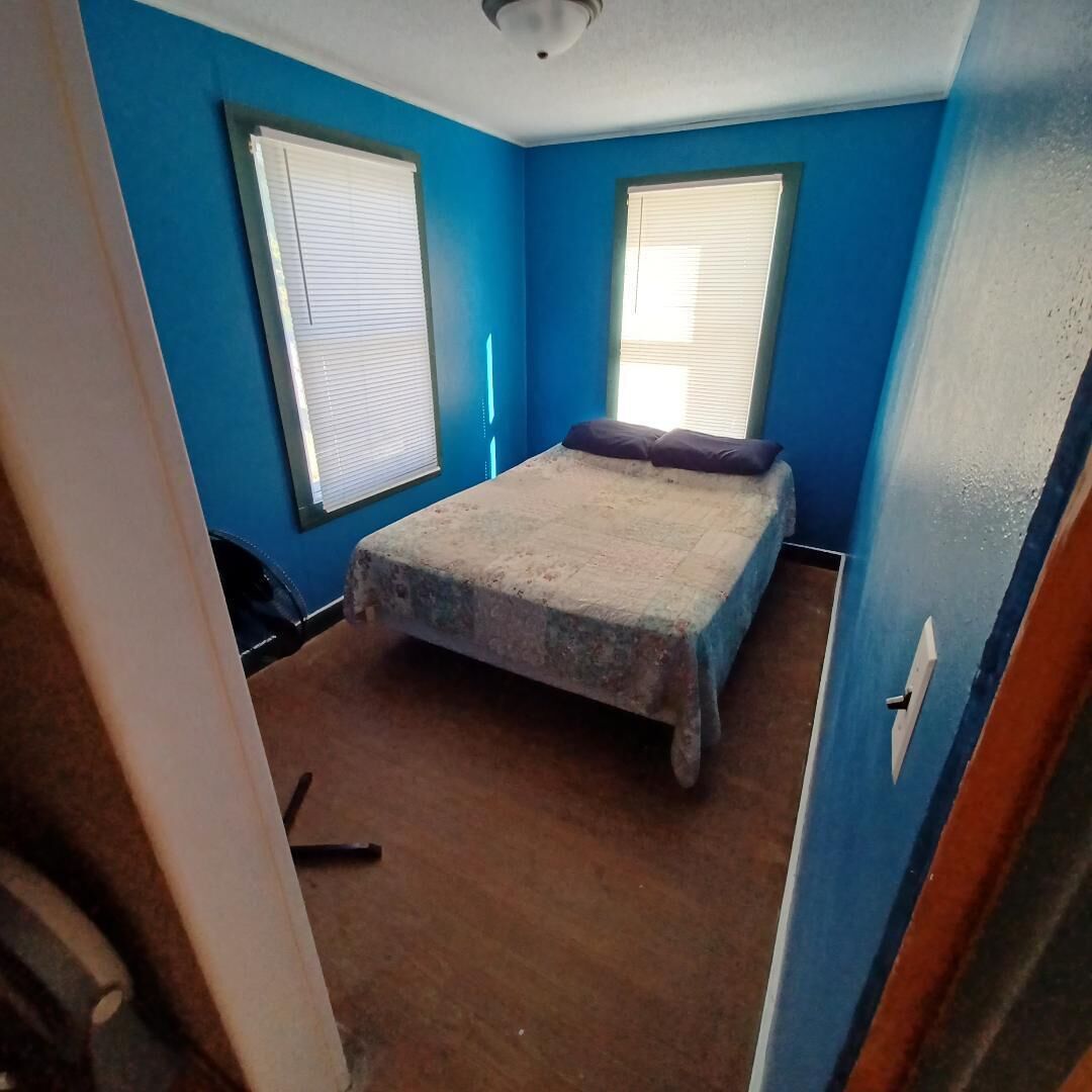 Ein Zimmer mit blauen Wänden, einem Bett mit weißer Matratze und einem blauen Kopfkissen, zwei Fenstern mit Jalousien und einem Holzboden.