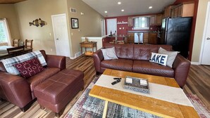 A living room with a brown leather sofa, a coffee table, and a kitchen in the background. - Beautiful lakefront, family-friendly! (Hustisford)