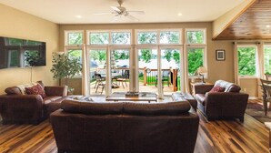 A spacious living room with a large sofa set, a coffee table, and a ceiling fan. There are large windows providing a view of the outdoors. - Beautiful lakefront, family-friendly! (Hustisford)