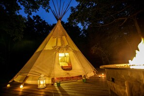 Une yourte avec un lit à l’intérieur, une lanterne et un foyer sur une terrasse en bois, le tout illuminé par le clair de lune.