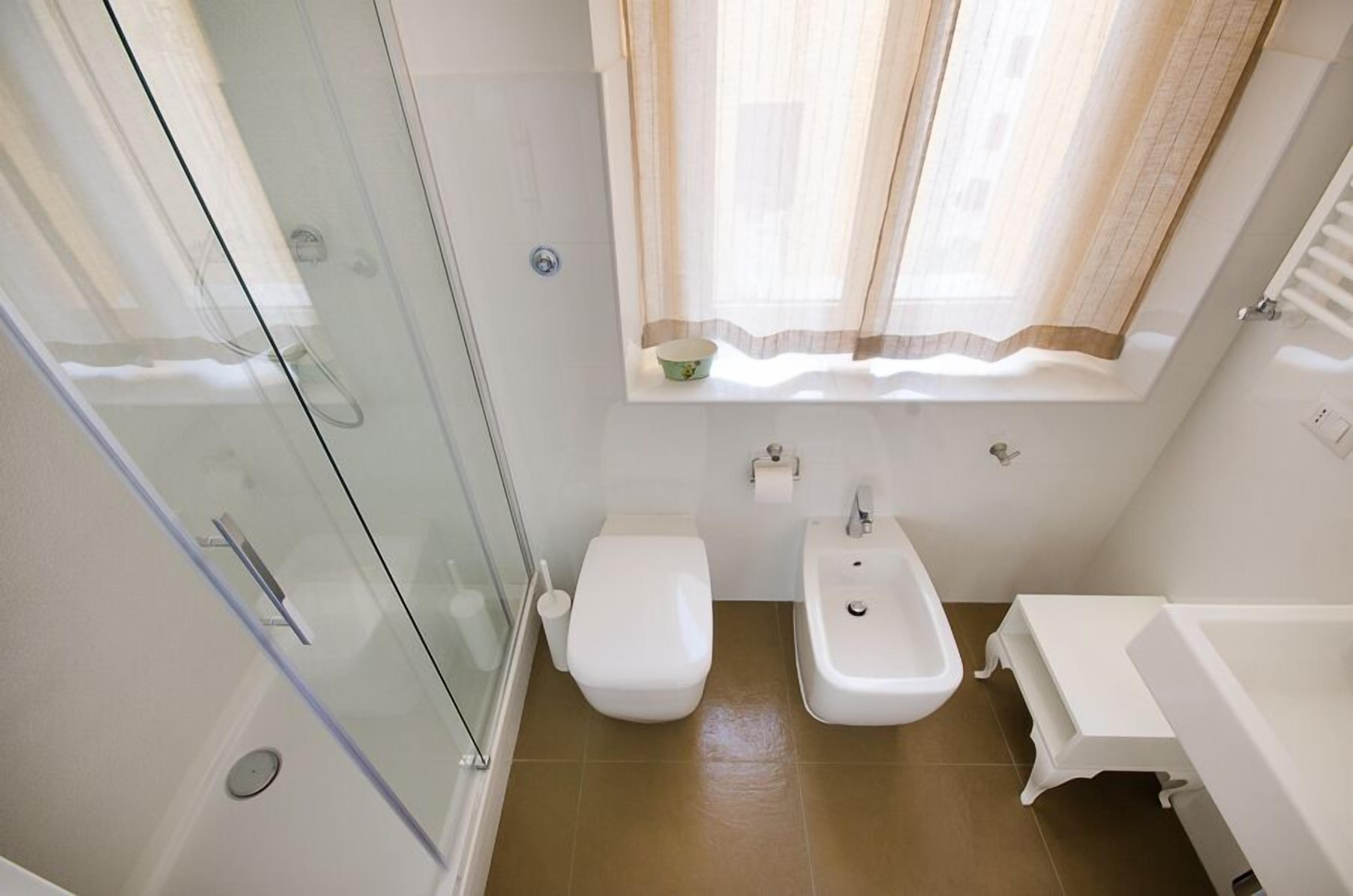 A compact bathroom with a glass shower enclosure, a toilet, a sink, and a window with curtains.