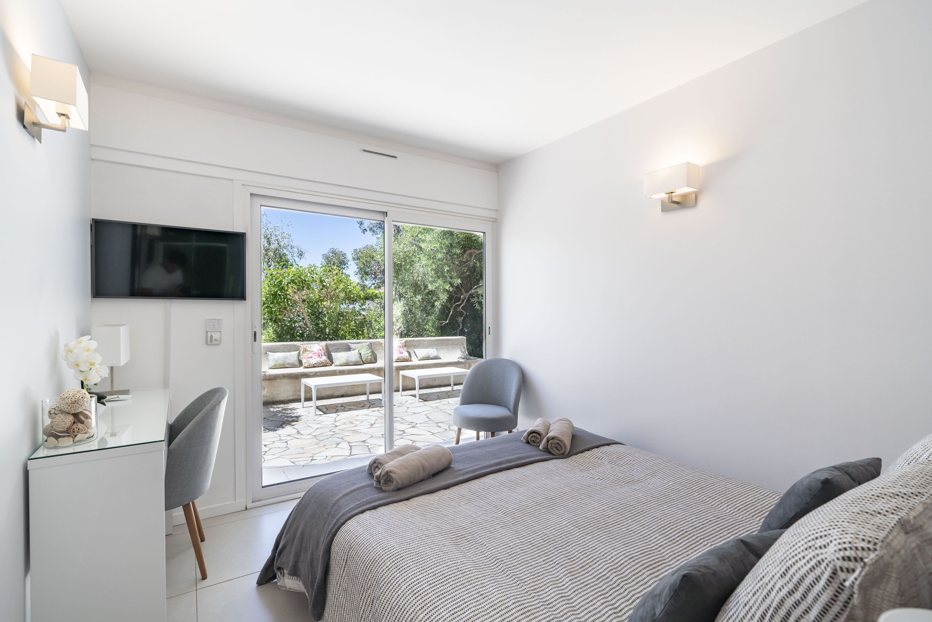 A modern bedroom with a large bed, a desk, and a chair. There is a sliding glass door leading to an outdoor seating area.