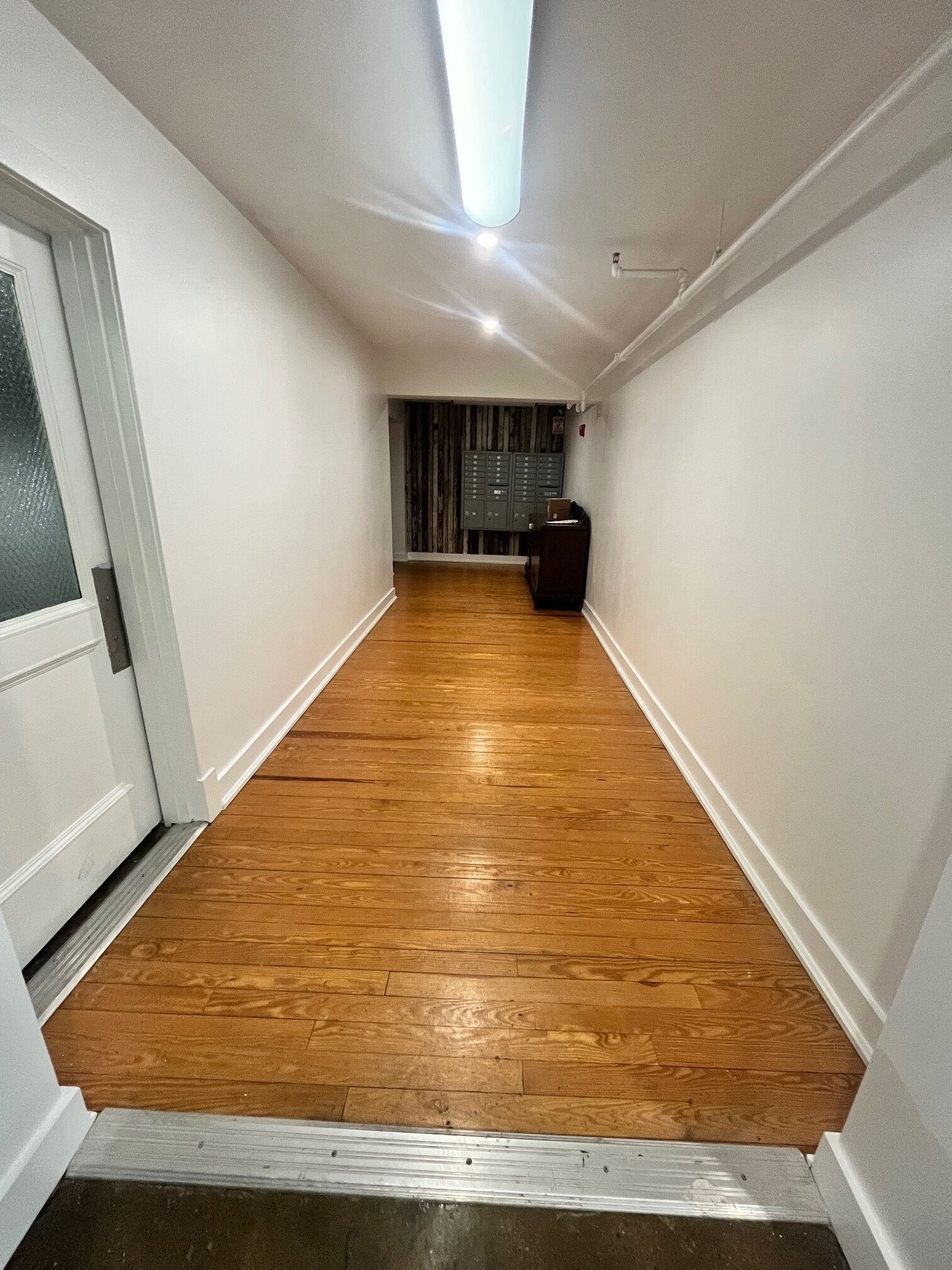A narrow hallway with wooden flooring, white walls, and a door on the left.