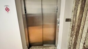 An elevator with a metallic finish, located in a hallway with wooden flooring and a textured wall to the right.