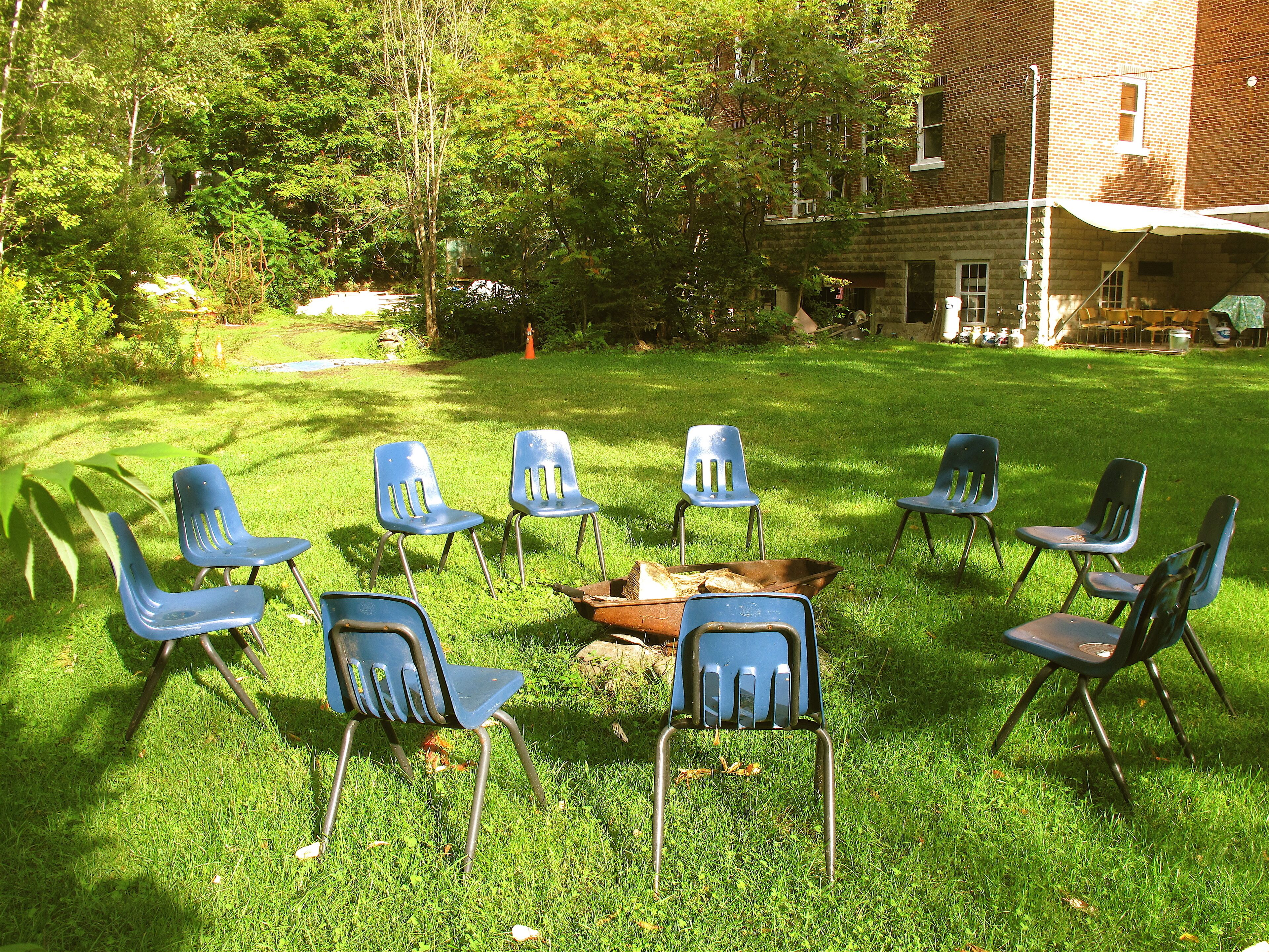 A grassy outdoor area with several blue plastic chairs arranged in a circle around a fire pit.