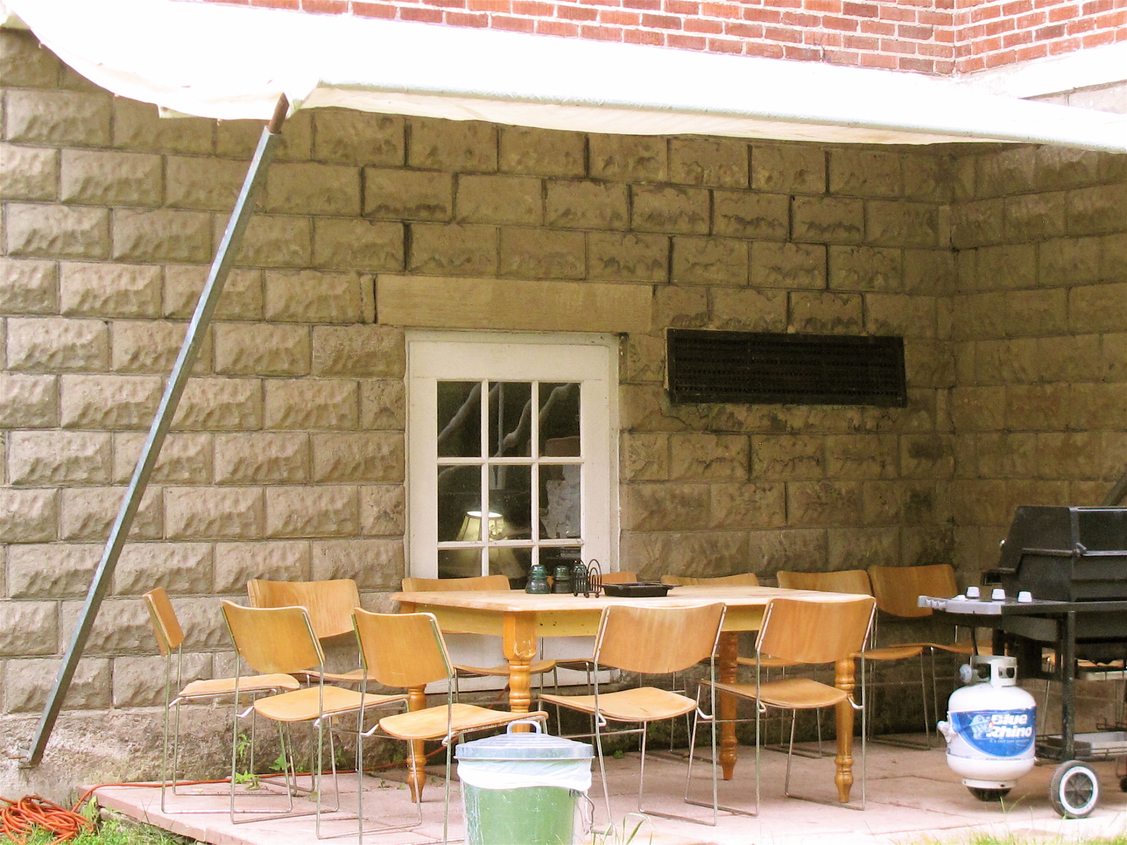 A stone patio with a wooden table and chairs, a window, and a propane tank.