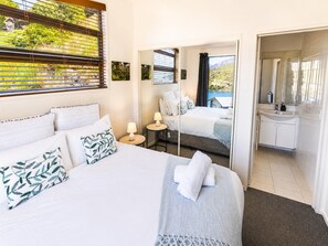 Een hotelkamer met een groot bed, een nachtkastje met lamp, een badkamer met wastafel en spiegel, en uitzicht op het landschap door het raam.