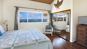 Une chambre avec un grand lit, une vue sur l’océan, un balcon avec un fauteuil et un téléviseur posé sur une commode en bois.