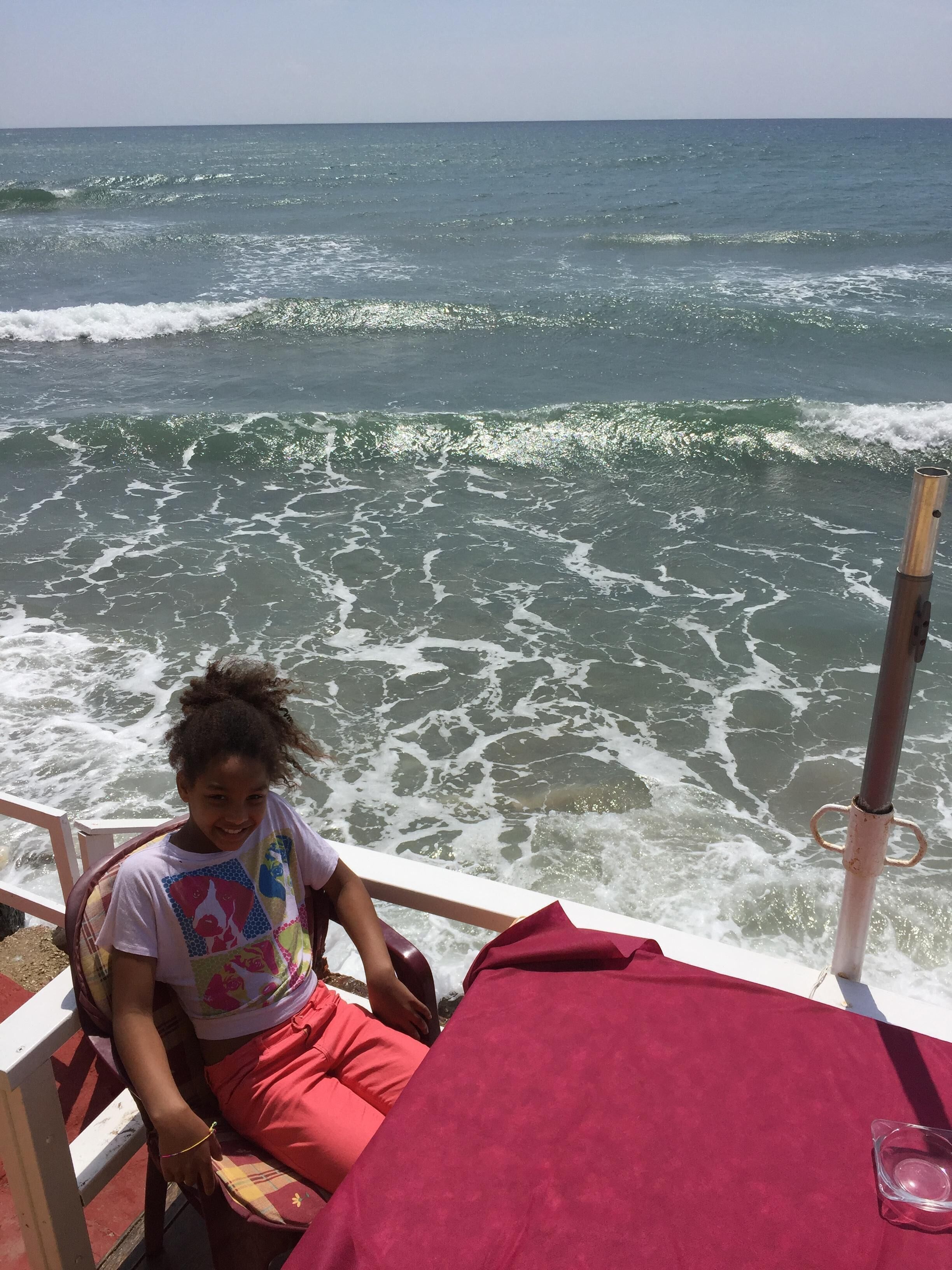 Eine Person sitzt an einem Stuhl am Meer, im Hintergrund brechen Wellen.