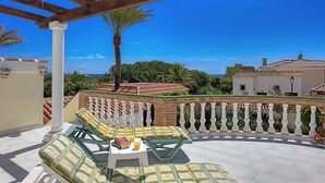 A covered outdoor seating area with a view of the sea, a palm tree, and a building with a tiled roof.