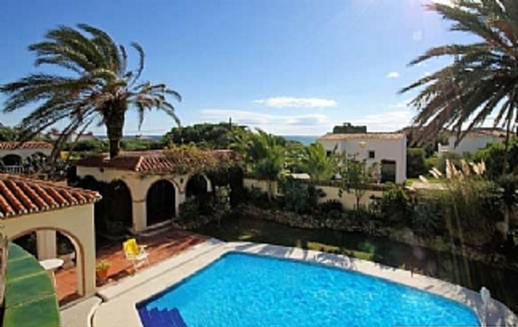 A swimming pool with a view of a coastal area, surrounded by buildings with arched doorways and palm trees.