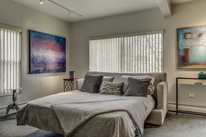 Een moderne slaapkamer met een groot bed, een nachtkastje, een raam met zonwering en twee abstracte schilderijen aan de wanden.