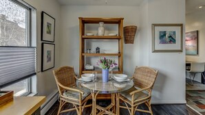 Un comedor con una mesa redonda de vidrio, sillas de mimbre, un jarrón azul con flores y una estantería de madera con objetos decorativos.
