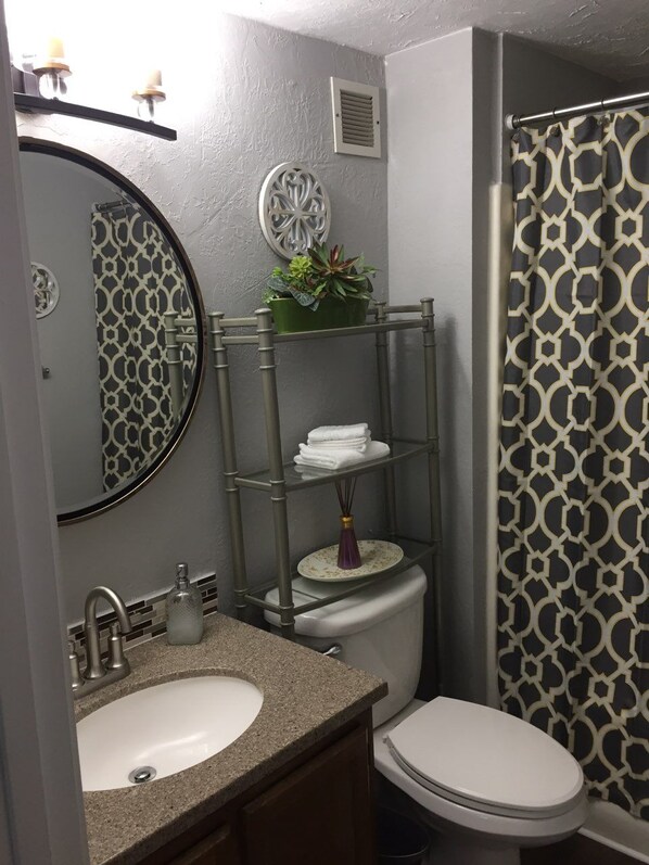 A bathroom with a round mirror, a sink, a toilet, a shower curtain, and a shelf with towels and a plant.