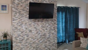 Una sala de estar con una pared de diseño de piedra, un televisor de pantalla plana montado arriba, una mesa de centro blanca, un sofá y una cortina azul.