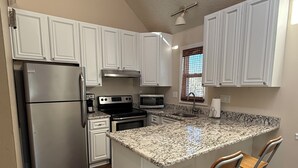 Una cocina con armarios blancos, electrodomésticos de acero inoxidable, encimera de granito y una ventana con persianas.