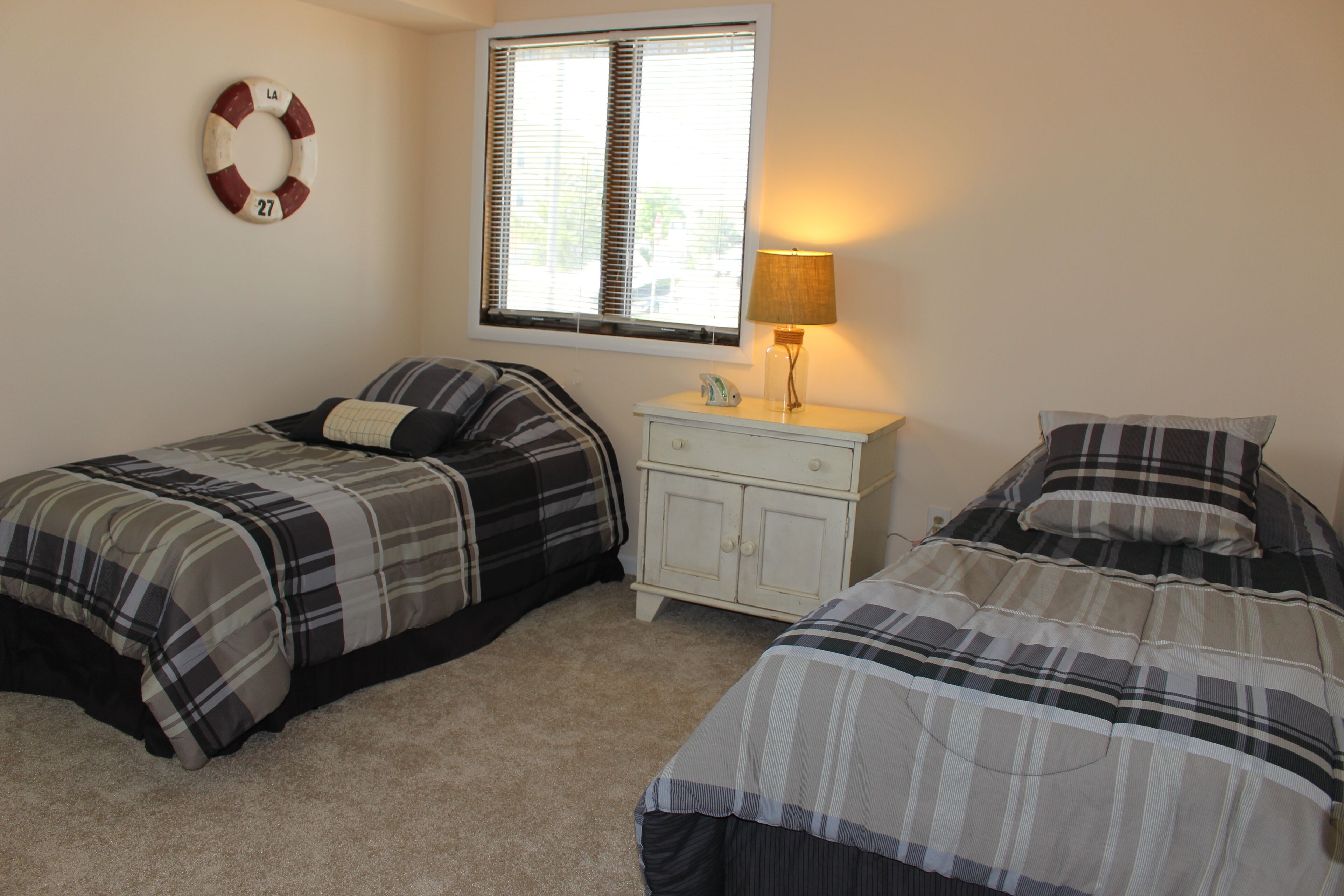 A bedroom with two beds, a nightstand with a lamp, a life preserver on the wall, and a window with blinds.