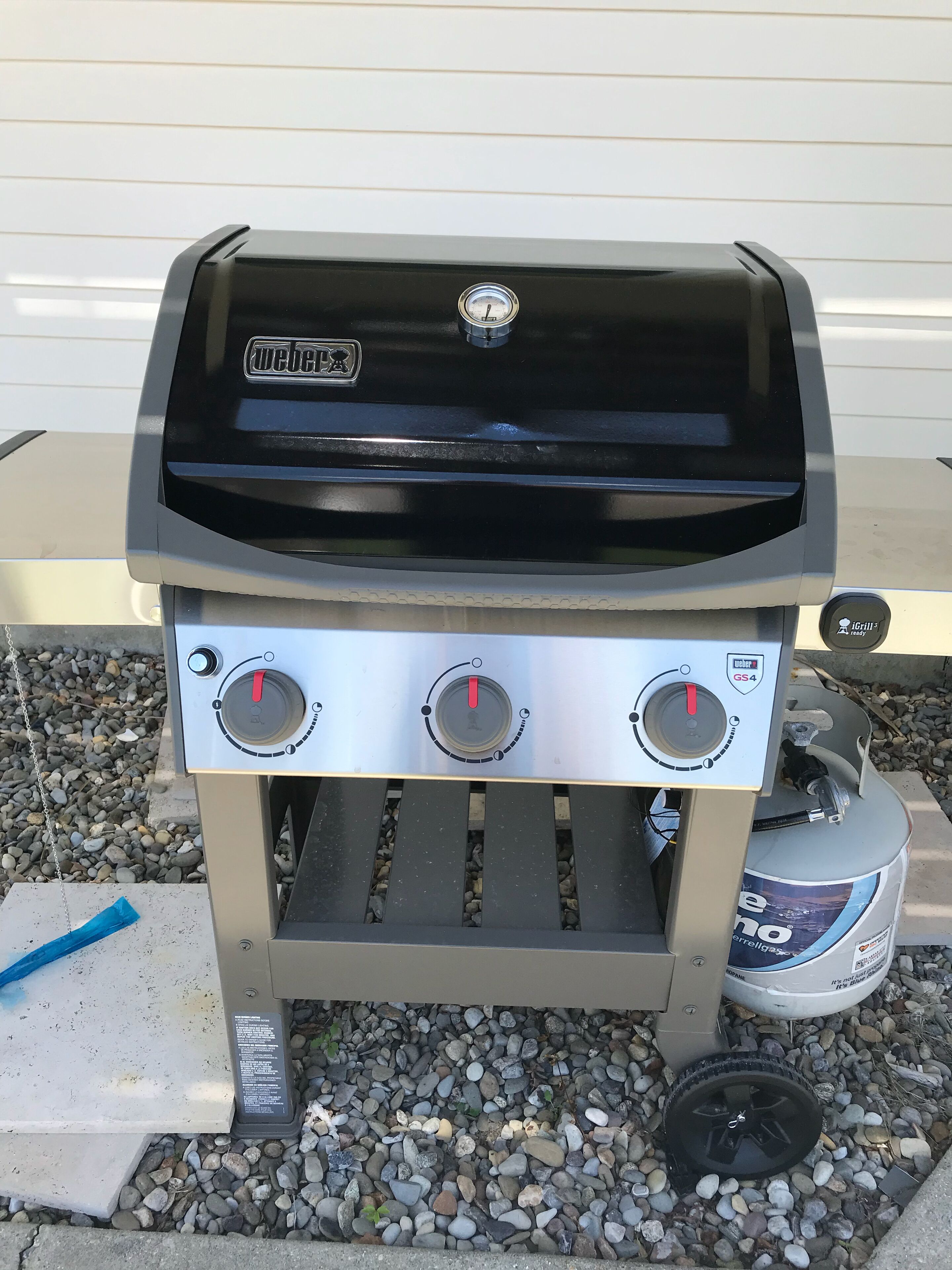 Una parrilla de gas Weber GS4 con tapa negra y tres perillas de control, ubicada sobre grava con un tanque de propano cerca.
