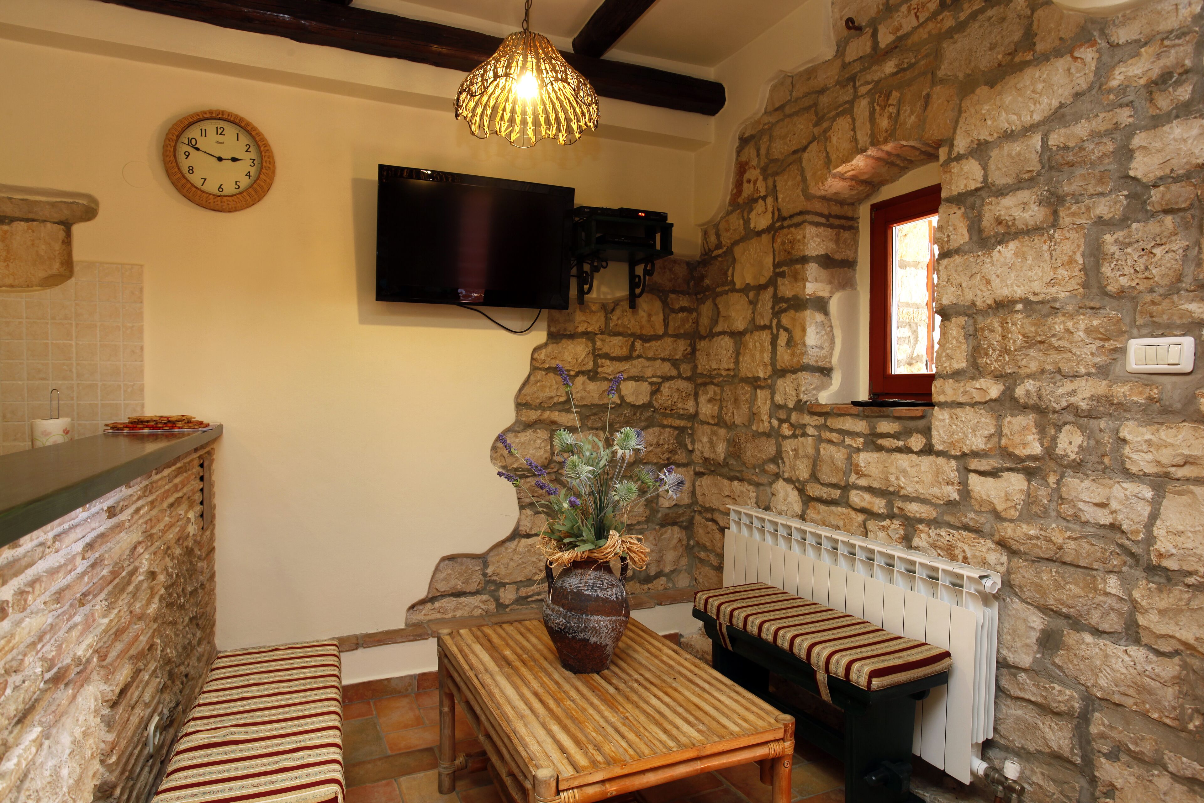 A stone-walled room with a wooden bench, a table with a vase, a clock, and a mounted TV.