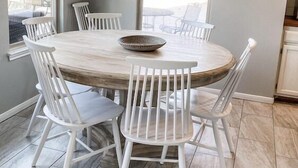 Un comedor con una mesa redonda de madera, sillas blancas, una lámpara colgante y un cartel enmarcado en la pared.