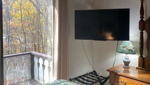 Une chambre à coucher avec un lit, un téléviseur fixé au mur, une lampe sur une table de chevet et une fenêtre donnant sur des arbres.