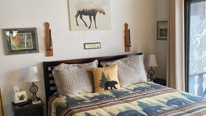 Une chambre avec un plafond en bois, un lit recouvert d’une courtepointe à motifs forestiers, une table de chevet avec une lampe, un tableau encadré représentant un élan, et une fenêtre donnant sur la forêt.