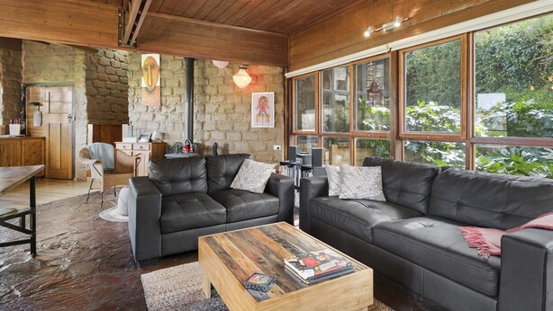 A cozy living room with a stone wall, wooden ceiling, and large windows overlooking greenery. - Driftaway Cottage - An enchanting escape (Ocean Grove)