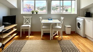 Une pièce de taille modeste comprenant une table à manger, des chaises, un téléviseur, un lave-linge et deux fenêtres équipées de stores.