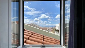 Une fenêtre donnant sur des toits couverts de tuiles en terre cuite et sur un ciel bleu dégagé parsemé de nuages.