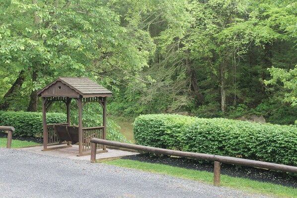 A wooden gazebo with a bench, surrounded by lush greenery and a gravel path.