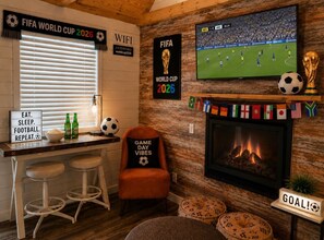 Una habitación con un cartel del Mundial de Fútbol 2026, un televisor mostrando un partido de fútbol, una chimenea y una zona de bar con taburetes y un cartel que dice '¡GOAL!'.